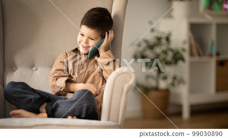 Glad caucasian little kid in casual calls by phone, talk, sits in armchair at spare time in living room interior Glad caucasian little kid in casual calls by phone, talk, sits in armchair at spare time in living room interior 99030890