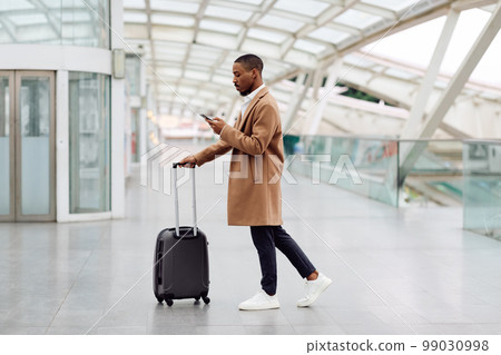 Online Check-In. Black Man Walking With Suitcase In Airport And Using Smartphone 99030998