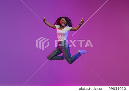 Cheerful Black Woman Jumping In Neon Light And Showing Thumbs Up 99031010