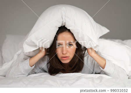 Tired sad caucasian millennial female lies on bed under white blanket, covers head with pillow, suffers from insomnia Tired sad caucasian millennial female lies on bed under white blanket, covers head with pillow, suffers from insomnia 99031032