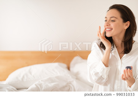 Cheerful caucasian millennial woman sitting on bed, applying moisturizing lipstick on lips, enjoy good morning 99031033