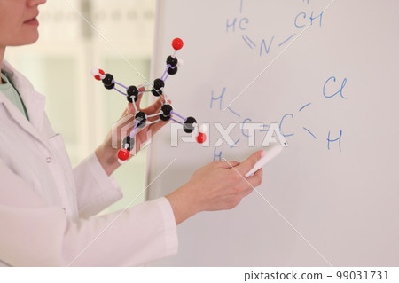 Female chemist holding molecular model and drawing chemical formulas on whiteboard. Female chemist holding molecular model and drawing chemical formulas on whiteboard. 99031731