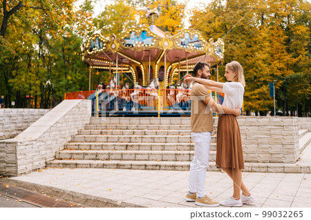 Wide shot of romantic young couple in love standing hugging looking at each other in amusement park in summer day.. Handsome bearded man and pretty blonde woman embracing enjoying weekend outdoors. 99032265