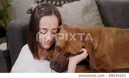 Young woman pets her dog while relaxing in the living room Young woman pets her dog while relaxing in the living room 99034201