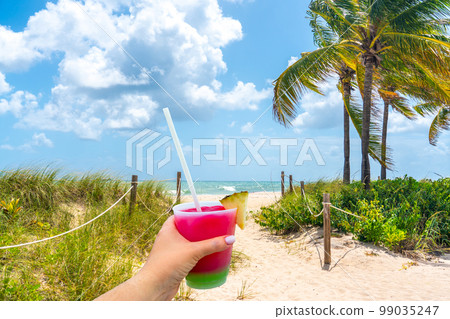 Hand holding a glass with cocktail and straw and Sand Path going to the ocean in Miami Beach, Florida Hand holding a glass with cocktail and straw and Sand Path going to the ocean in Miami Beach, Florida 99035247