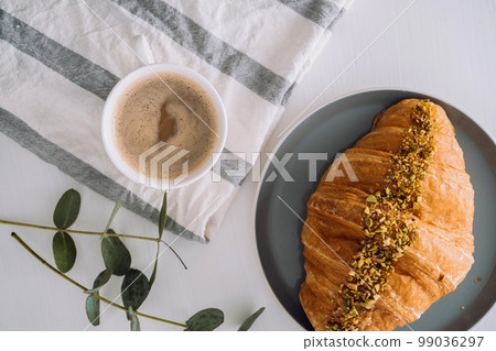 Flat lay of cup of cappuccino with kitchen towel, croissant and eucalyptus branch on the table 99036297