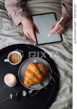 Woman using digital tablet for drawing while lying on bed and enjoying croissant with cappuccino Woman using digital tablet for drawing while lying on bed and enjoying croissant with cappuccino 99036307