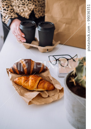 Delicious brown and chocolate croissants and coffee cups, woman having lunch at workplace, food delivery concept Delicious brown and chocolate croissants and coffee cups, woman having lunch at workplace, food delivery concept 99036351