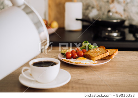 Homemade breakfast on the wooden table in the kitchen, blurred cup of coffee with kettle on the foreground 99036358