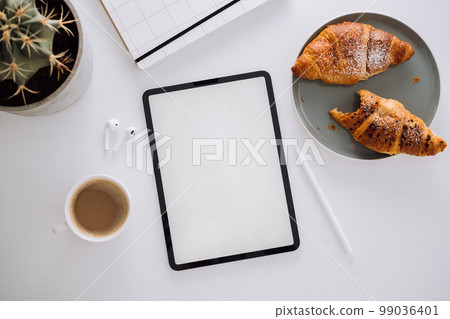 Flat lay of the workplace of the designer illustrator, digital tablet with white screen and stylus pen, cactus, coffee with croissants, notepad and earphones on the table 99036401