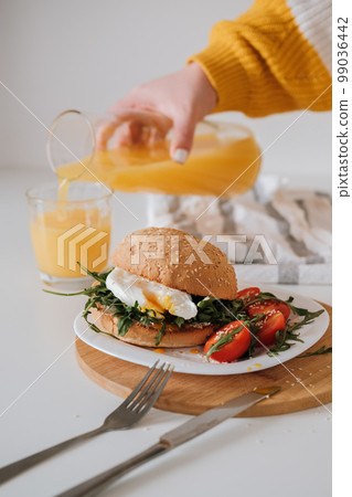 Breakfast sandwich with egg and arugula and cherry tomatoes on the table, woman pours orange juice into a glass 99036442