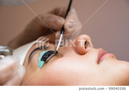 Close-up portrait of a woman on eyelash lamination procedure. The master applies tint to the eyelashes. Close-up portrait of a woman on eyelash lamination procedure. The master applies tint to the eyelashes. 99040386