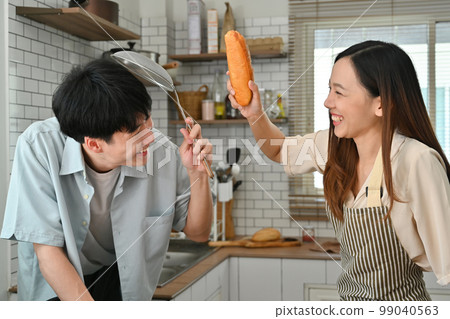 Overjoyed married couple using kitchenware and bread baguettes fighting in modern kitchen 99040563