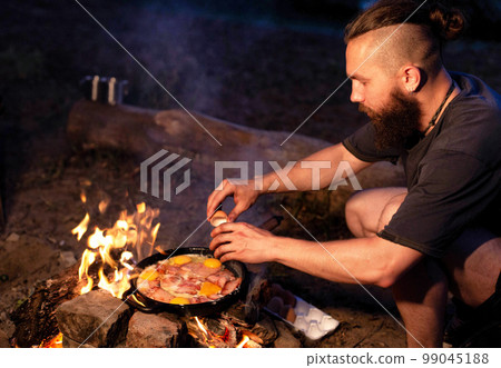 A young man with a beard prepares a dish of bacon with scrambled eggs on a fire. Cooking and outdoor recreation in summer. 99045188