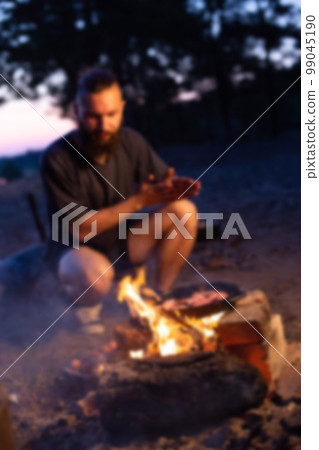 A young man with a beard prepares a dish of bacon with scrambled eggs on a fire. Cooking and outdoor recreation in summer, blurry A young man with a beard prepares a dish of bacon with scrambled eggs on a fire. Cooking and outdoor recreation in summer, blurry 99045190
