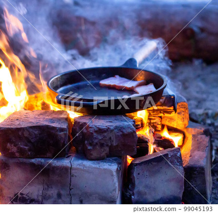 Frying bacon in a pan over a fire. Cooking outdoors in the evening. Frying bacon in a pan over a fire. Cooking outdoors in the evening. 99045193