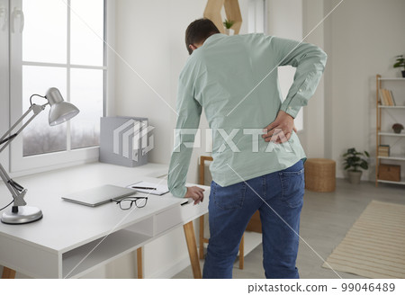 Adult man feels back pain after sedentary work at laptop, sitting at home in uncomfortable position on chair. 99046489