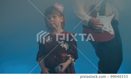 Little girl on stage in vintage dress, she sings into microphone and dances, her father plays an electric guitar. Color music is shining and smoke is billowing. Family time, creativity and hobbies Little girl on stage in vintage dress, she sings into microphone and dances, her father plays an electric guitar. Color music is shining and smoke is billowing. Family time, creativity and hobbies 99048333