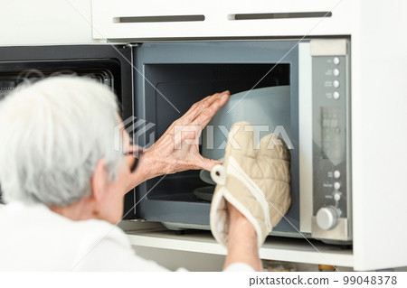 Asian senior woman wearing oven glove while taking food out of microwave oven,prevent damage and injury from heat accident,safety at home,hot bowl from cooking,old elderly heating food in kitchen 99048378