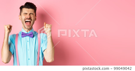 Relieved man shouting from happiness and joy, scream yes with closed eyes and clenched fists, celebrating victory, achieve goal and triumphing, standing over pink background 99049042