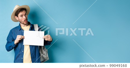 Excited tourist in straw hat, hitchhiking, showing blank piece of paper and looking amused, standing on blue background 99049168