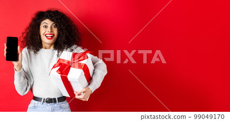 Valentines and lovers day. Excited smiling woman with curly dark hair, showing smartphone empty screen and holding surprise gift on holiday, showing online promo, red background 99049170