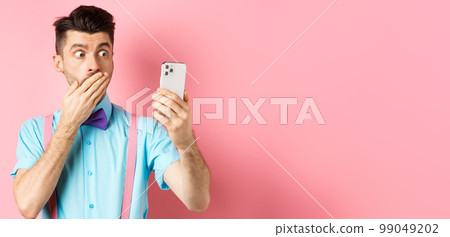 Technology concept. Silly young man looking shocked at smartphone screen while having video chat, standing on pink background and cover mouth with hand 99049202