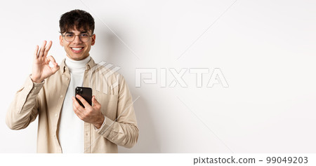 Technology and online shopping concept. Portrait of handsome modern guy in glasses showing OK gesture using smartphone, recommending app or shop, white background 99049203