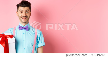 Holidays and celebration concept. Cheerful guy in bow-tie and suspenders bring gift box to party, holding present and smiling, standing over pink background 99049265