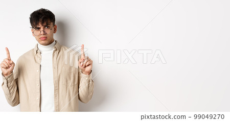 Portrait of young male model in glasses looking suspicious and frowning, pointing fingers up with disbelief, standing on white background Portrait of young male model in glasses looking suspicious and frowning, pointing fingers up with disbelief, standing on white background 99049270