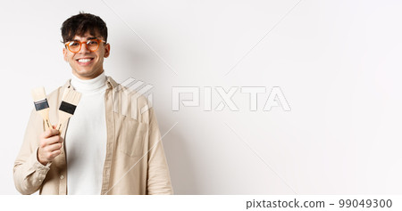 Hobbies and leisure concept. Stylish young man in glasses showing painting brushes and smiling, inviting to paint something, standing on white background 99049300