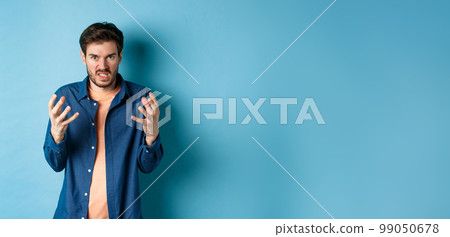 Annoyed and angry man raising hands and clench teeth outraged, stare pissed-off at camera, going to kill someone, standing on blue background Annoyed and angry man raising hands and clench teeth outraged, stare pissed-off at camera, going to kill someone, standing on blue background 99050678