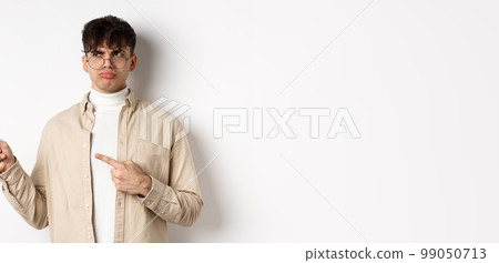Disappointed guy in glasses looking and pointing left, sulking displeased, complaining on bad offer, standing upset on white background 99050713