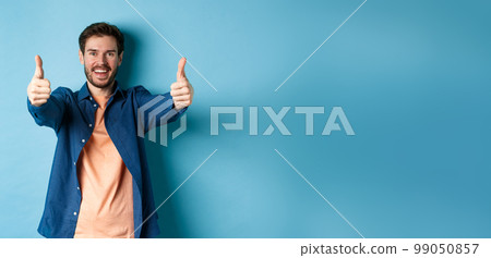 Handsome young man smiling and showing thumbs up in approval, say yes with happy face, standing on blue background Handsome young man smiling and showing thumbs up in approval, say yes with happy face, standing on blue background 99050857
