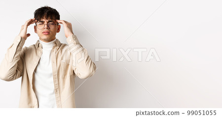 Image of young man in glasses having headache, touching head temples and frowning from painful migraine, feel dizzy, standing on white background Image of young man in glasses having headache, touching head temples and frowning from painful migraine, feel dizzy, standing on white background 99051055