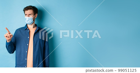 Covid-19 and healthcare concept. Confused and doubtful man in face mask frowning, pointing and looking at empty space, standing on blue background Covid-19 and healthcare concept. Confused and doubtful man in face mask frowning, pointing and looking at empty space, standing on blue background 99051125