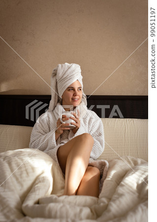 A woman sits on a bed wearing a white dressing gown, with a towel covering her head and hold a cup of warm drink A woman sits on a bed wearing a white dressing gown, with a towel covering her head and hold a cup of warm drink 99051197