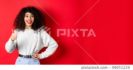 Happy young woman with curly hair praising good work, say well done and show thumb up gesture, approve and praise you, standing on red background Happy young woman with curly hair praising good work, say well done and show thumb up gesture, approve and praise you, standing on red background 99051229