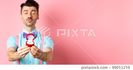 Valentines day. Silly boyfriend in bow-tie pucker lips, close eyes and waiting for kiss after making proposal, showing golden engagement ring in small red box, standing on pink background Valentines day. Silly boyfriend in bow-tie pucker lips, close eyes and waiting for kiss after making proposal, showing golden engagement ring in small red box, standing on pink background 99051259