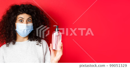 Covid-19, pandemic and quarantine concept. Excited girl with curly hair, wearing medical mask, showing bottle of hand sanitizer or antiseptic, standing on red background 99051329