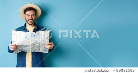 Cheerful tourist in summer hat planning travel route on vacation, looking at sighseeing map and smiling excited, standing on blue background 99051592