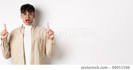 Amazed and excited man in glasses showing awesome promo deal, pointing fingers up and open mouth in awe, standing against white background 99051598