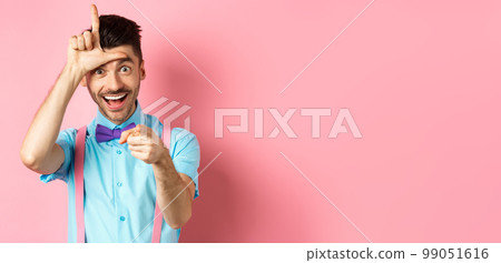 Image of funny guy pointing at you camera and showing loser sign, mocking person who lose, standing over pink background 99051616
