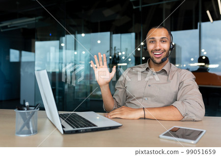 Portrait of tech support and online customer service worker, man with video call headset smiling and looking at camera, african american man in shirt working inside office using laptop. Portrait of tech support and online customer service worker, man with video call headset smiling and looking at camera, african american man in shirt working inside office using laptop. 99051659