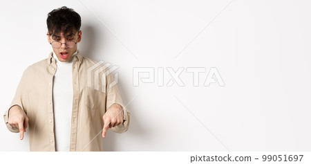 Amazed young guy in glasses look and point down, open mouth and say wow impressed, checking out discount offer, standing on white background Amazed young guy in glasses look and point down, open mouth and say wow impressed, checking out discount offer, standing on white background 99051697