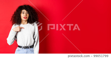 Surprised attractive female model pointing at herself, raising eyebrows and looking with disbelief at camera, standing on red background 99051799