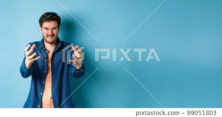 Angry tensed man wants to strangle someone, stretch out hands to choke enemy, biting lip and looking furious, standing on blue background Angry tensed man wants to strangle someone, stretch out hands to choke enemy, biting lip and looking furious, standing on blue background 99051803