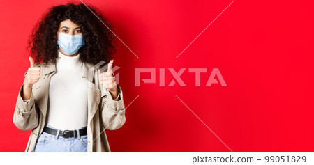 Covid-19, social distancing and quarantine concept. Cheerful stylish woman wearing trench coat and medical mask for going outside during pandemic, showing thumb up 99051829