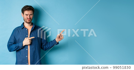 Disappointed young man frowning and pointing fingers right at empty space, looking upset and displeased, complaining, standing on blue background 99051830