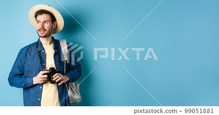 Handsome positive guy in summer hat, holding photo camera and looking aside, tourist taking pictures on vacation, standing with backpack on blue background Handsome positive guy in summer hat, holding photo camera and looking aside, tourist taking pictures on vacation, standing with backpack on blue background 99051881